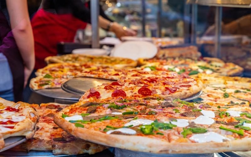 Pizzeria glass window. Variety of italian pizzas in a shop display, street food. Kitchen workers arranging the pizzas.